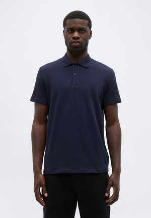 Giovane uomo in piedi di fronte, indossa una polo a maniche corte blu navy e pantaloni neri, su uno sfondo grigio chiaro uniforme.