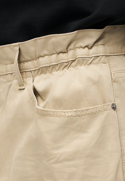 Pantalon beige avec taille élastique et coutures visibles, porté avec un haut noir rentré dedans.