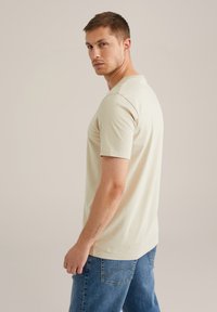 Bēšas krāsas īss piedurkņu T-krekls ar apaļu kakla izgriezumu, izgatavots no mīksta materiāla, kombinēts ar gaiši zilām džinsa biksēm. Redzamas smalkas šūšanas detaļas.