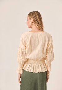 Blusa de manga abullonada en beige claro y texturizada con cintura fruncida, combinada con una falda verde. Tela suave, diseño relajado y rayas sutiles.