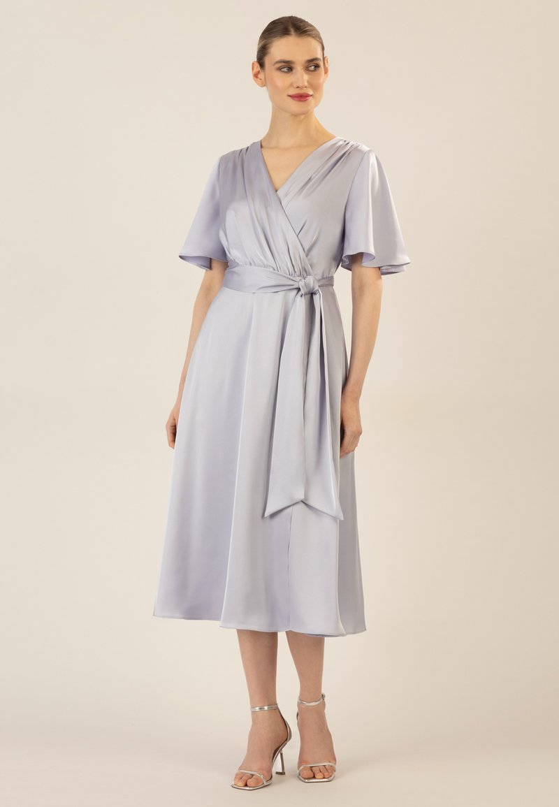 Helles Lavendel-Wickelkleid mit kurzen Volantärmeln, Taillenguertel und knielangem Saum aus glattem Stoff. Kombiniert mit silbernen Absatzsandalen.