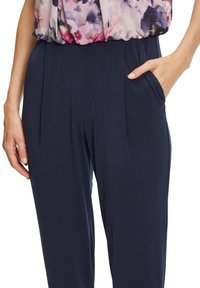 Femme portant un pantalon taille haute bleu marine avec des poches latérales, associé à un chemisier floral rose et violet rentré à l'intérieur.