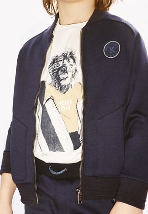 Marineblauwe bomberjack met een ritssluiting aan de voorkant en geribbelde manchetten, gedragen over een crème T-shirt met een leeuwengrafiek en de tekst "FRENCH LOVER".