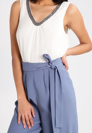 Haut sans manches blanc avec un col argenté à motifs, associé à un pantalon taille haute bleu clair avec une ceinture nouée et des plis.