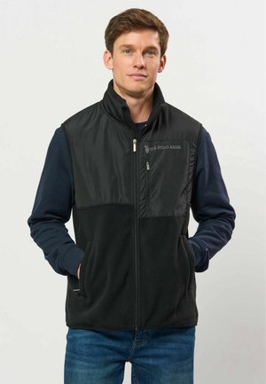 Homme portant un gilet noir zippé par-dessus une chemise bleu marine à manches longues, les mains dans les poches du gilet, debout devant un fond uni.