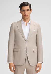 Beige Leinenanzug mit strukturiertem Muster, einreihige Jacke, mit Revers, und zwei Knöpfen; getragen über einem weißen Hemd mit Kragen.