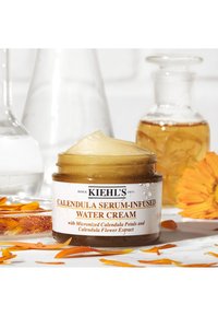 Kiehl's Calendula Serum-Infused Water Cream i en klar krukke med et gyllent lokk, fylt med et kremete, blekgult produkt og omgitt av blomstblader.