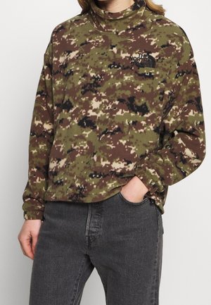 Person trägt einen North Face Fleece-Pullover mit grün-braunem Camouflage-Muster und hohem Kragen sowie schwarze Jeans, Hand in der Tasche.