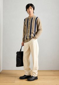 Beige och marinblå randig knapp-up cardigan, krämfärgade byxor och svarta slip-on skor. Modellen håller en svart tote-väska. Bakgrund av trägolv.