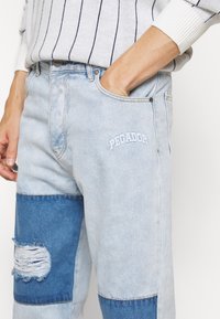 Vaqueros de mezclilla azul claro con una mezcla de parches en azul claro y oscuro, área de rodilla desgastada y "PEGADOR" bordado en la parte delantera.