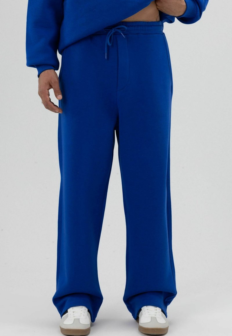 Blauwe sweatpants gemaakt van zachte stof, met een trekkoord in de taille, een brede pijp en een gladde textuur. Geaccentueerd met witte schoenen.