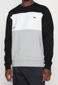 Uomo che indossa una felpa Lacoste a blocchi di colore nero, bianco e grigio con un piccolo logo di coccodrillo verde sul petto.