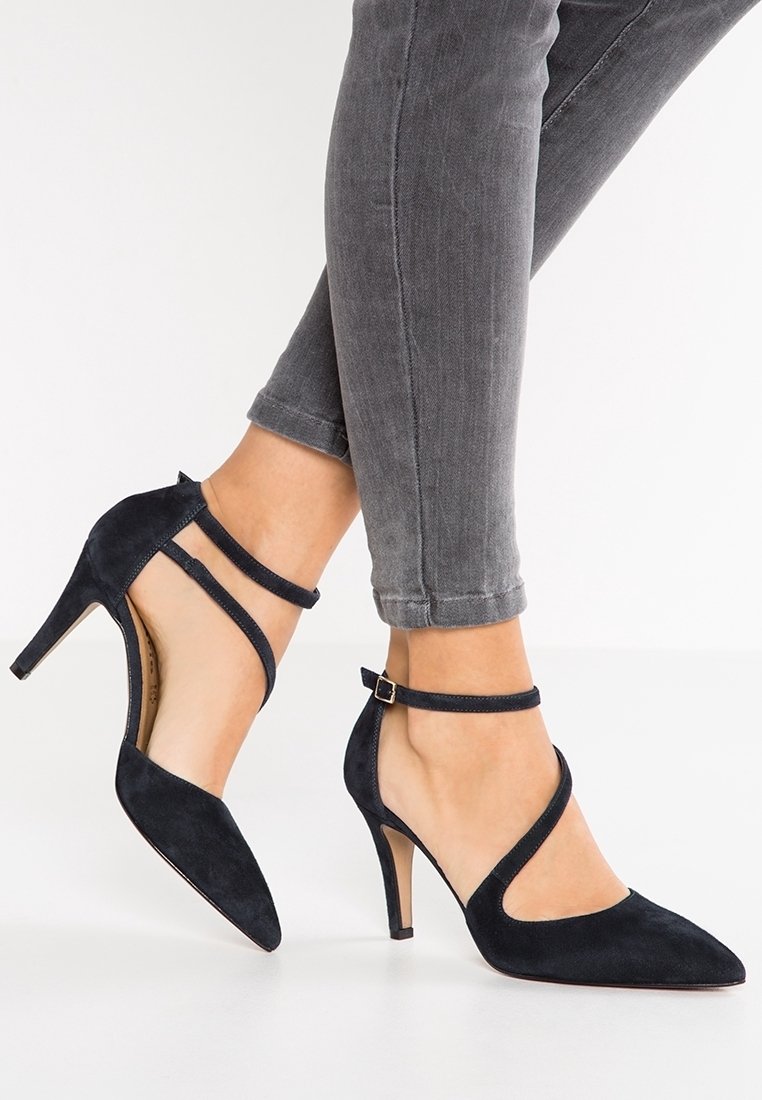 Femme portant des talons aiguilles en daim noir à bout pointu avec doubles brides à la cheville et jean skinny gris sur fond blanc.