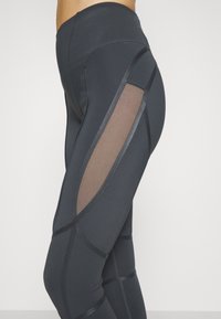 Spodnie legginsy z wysokim stanem w ciemnoszarym kolorze, z przezroczystymi panelami siateczkowymi po bokach, teksturowanym materiałem oraz kontrastowymi detalami szwów.