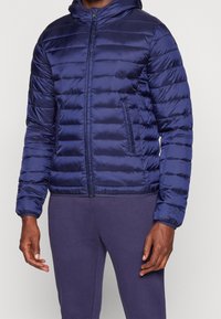 Marine pufferjack met capuchon, geborduurd textuur, voorrits en zijzakken. Beschikbaar met elastische manchetten en een getailleerd ontwerp.