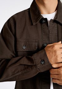 Camicia di denim marrone con colletto, tasca frontale e bottoni neri. Tessuto liscio con dettagli di cucitura visibili.