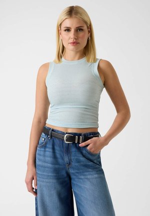 Blondynka ubrana w jasnoniebieski bezrękawnik typu crop top i wysokowciskane niebieskie dżinsy z czarnym paskiem, stojąca z jedną ręką w kieszeni.