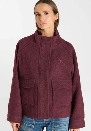 Donna che indossa una giacca di lana color bordeaux con collo alto, maniche lunghe, due grandi tasche frontali e jeans blu.