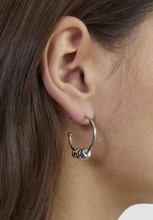 Nærbillede af et venstre øre med brunt hår, der bærer en sølv hoop-ørering med tre små ringe på undersiden.