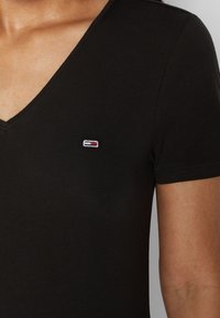 Närbild av en svart V-ringad t-shirt med en liten broderad logotyp på vänster bröst, som framhäver tygtexturen och halslinjen.