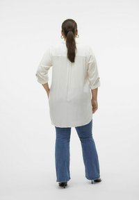 Lang hvid bluse med trekvartærmer, der har en knapdetalje ved manchetterne, parret med blå flagrende jeans. Glat tekstur.