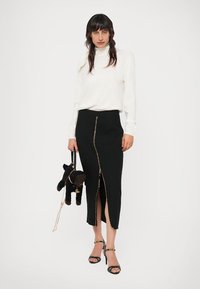 Sieviete, tērpta baltā turtleneckā un melnā svārku ar priekšējo rāvējslēdzēju, tur melnu somiņu suns formas, valkājot melnas augstpapēžu sandalītes.