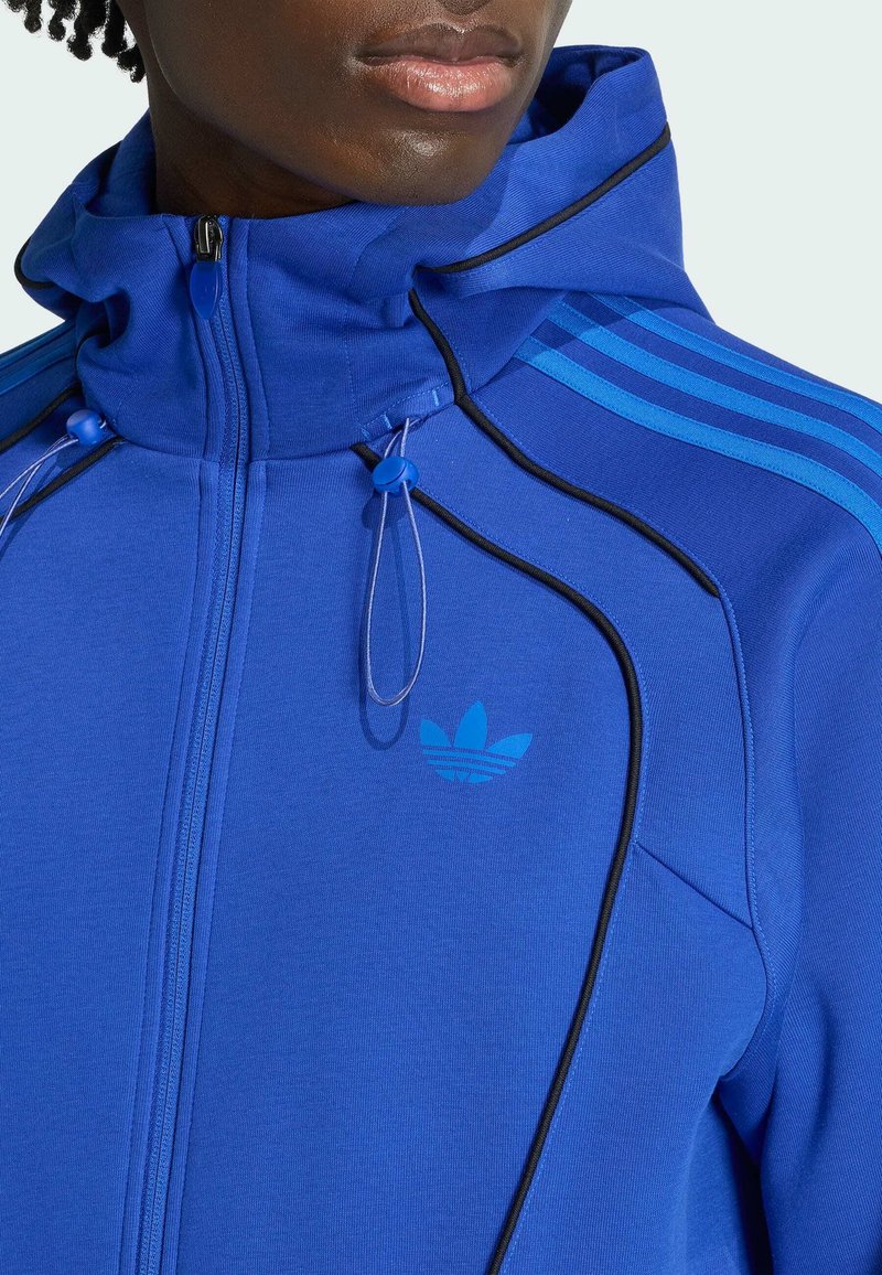 Gros plan d'une personne portant un sweat à capuche zippé bleu avec col montant, passepoil noir, cordon de serrage, et logo Adidas ainsi que des bandes sur l'épaule.