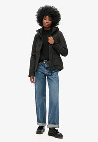 Schwarze isolierte Jacke mit Kapuze, frontalen Taschen und Reißverschlussdetails. Getragen über einem schwarzen Pullover, kombiniert mit hellblauen Schlagjeans und schwarzen Schuhen.