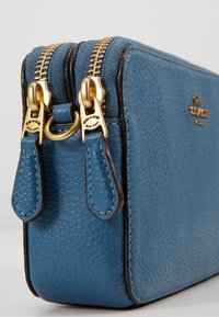 Blue leather rectangular pouch with double golden zipper, embroidered logo, and smooth texture. Accented with dark edges and zipper pulls.