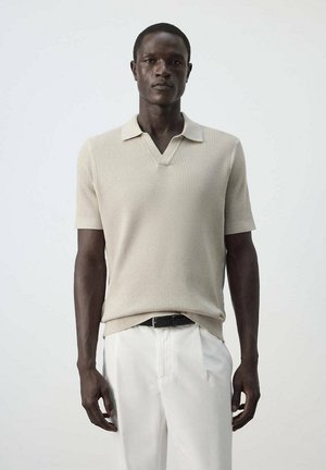Homme portant un polo en maille beige à manches courtes, rentré dans un pantalon blanc avec une ceinture noire, debout devant un fond uni.