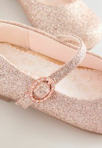 Mirdzoši rozā baleta kurpes ar siksniņu un dekoratīvu rozā zelta sprādzi, ar mīkstu iekšējo apdari un teksturētu mirdzošu ārējo virsmu.