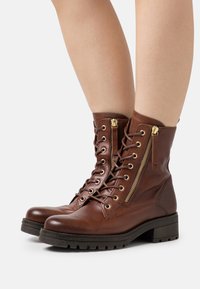 Botas de piel marrón hasta el tobillo con acentos de cremallera dorada, parte delantera con cordones, suelas acolchadas y diseño texturizado para mayor agarre.