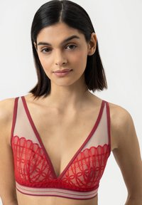 Jonge vrouw met stijlvol donker haar, gekleed in een rode kant bralette met gaaspanelen en een gestreepte elastische band, tegen een plain witte achtergrond.