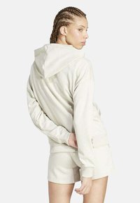 Donna con i capelli intrecciati, che indossa un felpa color crema e pantaloni corti abbinati, posando con una mano dietro la schiena e guardando oltre la spalla.