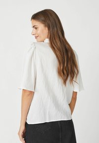 Blusa bianca a maniche corte con una texture stropicciata, decorata con un lieve volant al collo e una vestibilità comoda. Indossata con una gonna scura.
