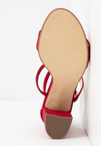 Sandale à talons hauts en suède rouge avec un bout carré et de fines sangles à la cheville. La semelle est beige avec un motif texturé pour une meilleure adhérence.