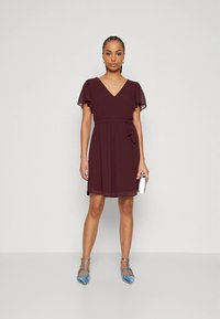 Vīnogu sarkana īsa kleita ar plūstošām piedurknēm, V veida izgriezumu, jostu viduklī un daļēji caurspīdīgu audumu. Valkāta ar gaiši zilām punktveida dzīvokļa stilā kurpēm.