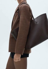 Ruskea houndstooth-kuviollinen bleiseri, jossa kolme nappikoristetta, yhdistettynä suureen tummanruskeaan nahkalaan laukkuun, jossa on sileä pinta ja olkahihnat.