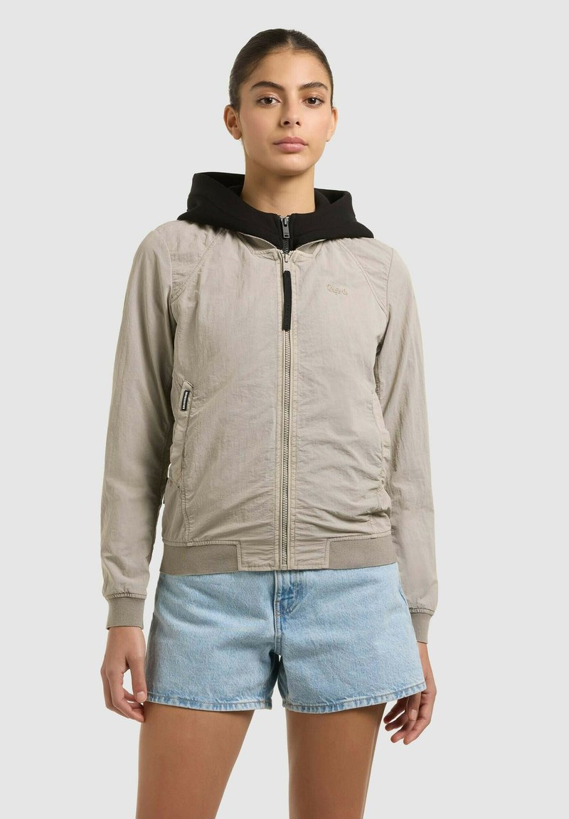 Jeune femme aux cheveux foncés tirés en arrière porte une veste beige à fermeture éclair sur une couche noire à capuche et un short en denim bleu clair.