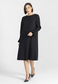 Czarna sukienka w kształcie litery A z lekkiego materiału, z długimi rękawami z akcentami, okrągłym dekoltem i długością do kolan.