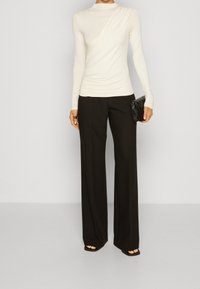 Långärmad krämfärgad topp med draperad design, kombinerad med högmidjade svarta vida byxor. Accentuera med en liten svart clutchtvätt.