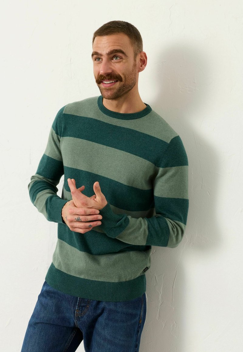 Mann mit kurzen Haaren und Schnurrbart, der einen grün gestreiften Pullover und blaue Jeans trägt und vor einer weißen, strukturierten Wand steht.