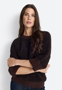 Pullover bulky di colore marrone scuro con tessuto strutturato, polsini e orlo a costine, vestibilità rilassata, indossato sopra un paio di jeans con le braccia incrociate.