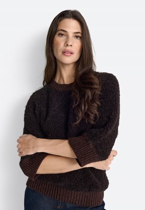 Pull sombre en brun foncé avec un tissu texturé, des poignets et un ourlet côtelés, coupe décontractée, porté sur un jean avec les bras croisés.