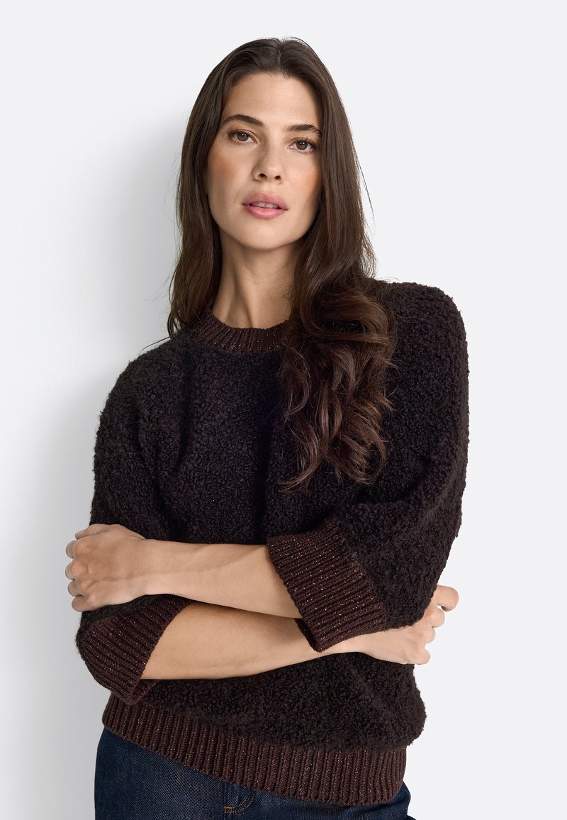 Pullover bulky di colore marrone scuro con tessuto strutturato, polsini e orlo a costine, vestibilità rilassata, indossato sopra un paio di jeans con le braccia incrociate.