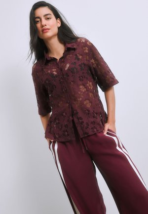 Femme portant une chemise en dentelle florale bordeaux transparente et un pantalon large assorti bordeaux avec des bandes blanches sur les côtés, debout devant un fond uni.