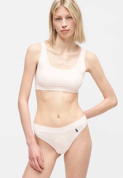 Młoda kobieta o krótkich blond włosach, ubrana w dopasowany jasnoróżowy sportowy biustonosz i majtki z małym ciemnym logo na pasku majtek.