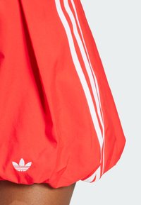 Červená atletická sukňa vyrobená z ľahkého materiálu, s bielymi pruhmi Adidas po stranách a malým bielym logom pri leme.