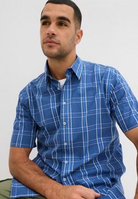 Camicia a maniche corte a quadri blu con linee bianche e azzurre, dotata di colletto e chiusura frontale con bottoni. Tessuto liscio, vestibilità su misura.