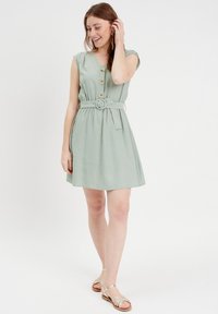 Robe courte vert menthe avec des manches courtes, des boutons sur le devant, une taille ceinturée et une jupe évasée. Portée avec des sandales métalliques à lanières.